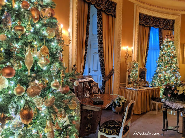 A bedroom decorated in gold, with a large Christmas tree on the left, another Christmas tree on the right, and a reflection of a tree in a vanity mirror