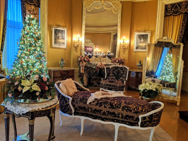 A bedroom decorated in gold, with purple furnishings. A Christmas tree on the left, and a Christmas tree reflected in a mirror on the right. 