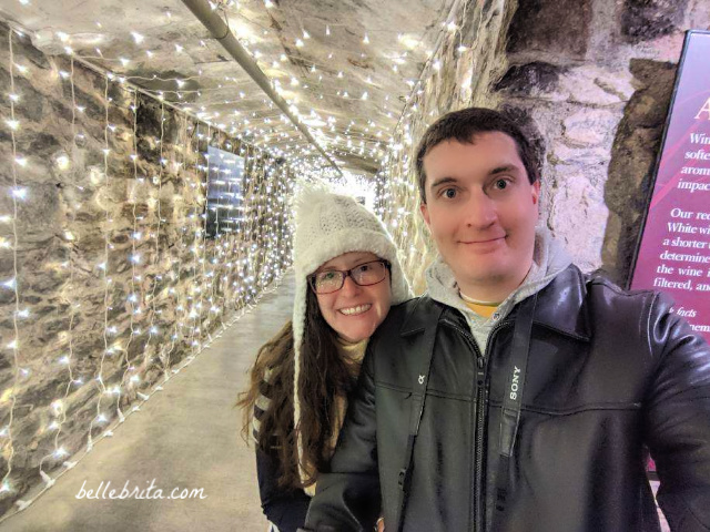 Woman and man smiling, standing in a long hallway decorated with twinkling lights. Winery at Biltmore.