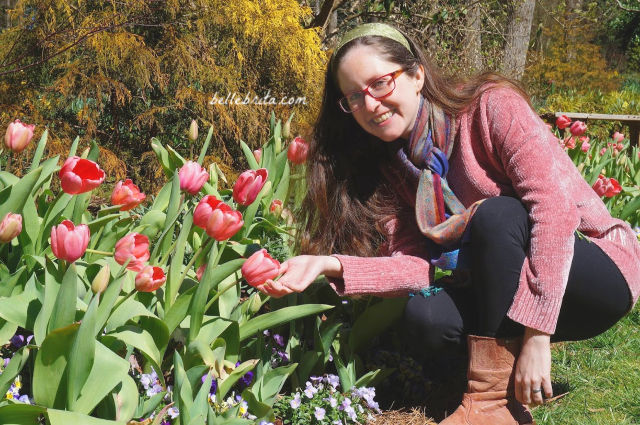 White woman wearing a pink Marley Lilly sweater and black leggings, crouched near pink tulips.