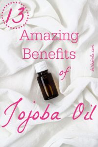 Amber glass bottle on sheets. Text overlay reads: "13 Amazing Benefits of Jojoba Oil"