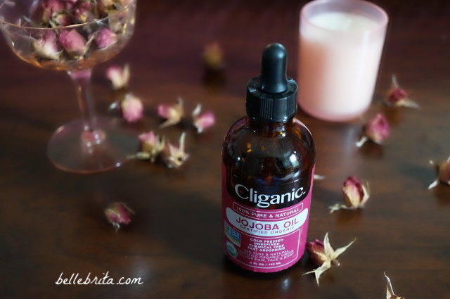 Bottle of jojoba oil on a wooden surface, with dried pink roses scattered around. 