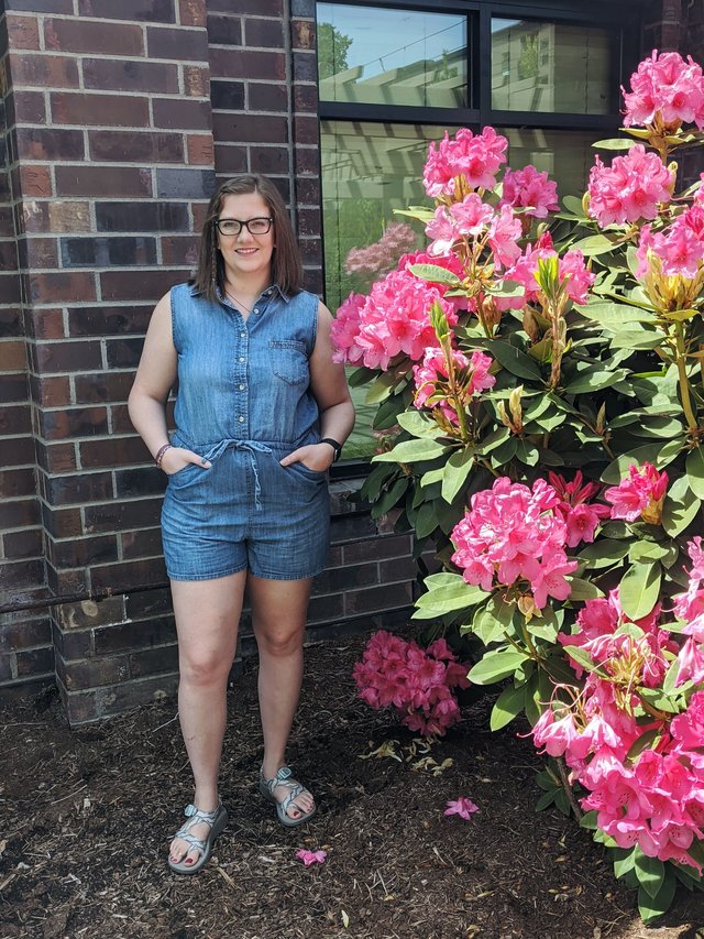 White woman wearing a denim romper standing by pink flowers