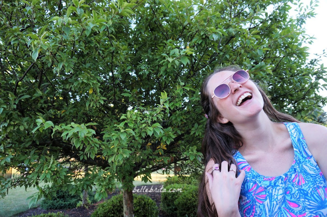 White woman wearing sunglasses and blue print dress standing in front of a tree