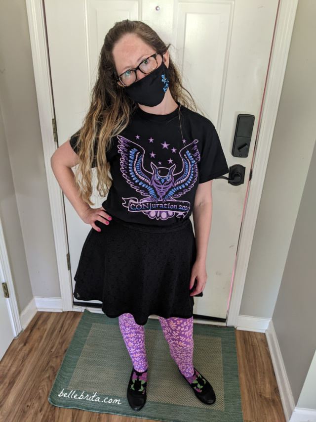 Full-body shot of white woman wearing a black face mask, a patterned black t-shirt, a black skirt, patterned pink and purple leggings, and black shoes, standing in a front hallway.