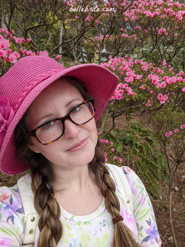 Close-up shot of white woman with two brown braids, a pink straw hat, and tortoiseshell glasses standing among pink flowers