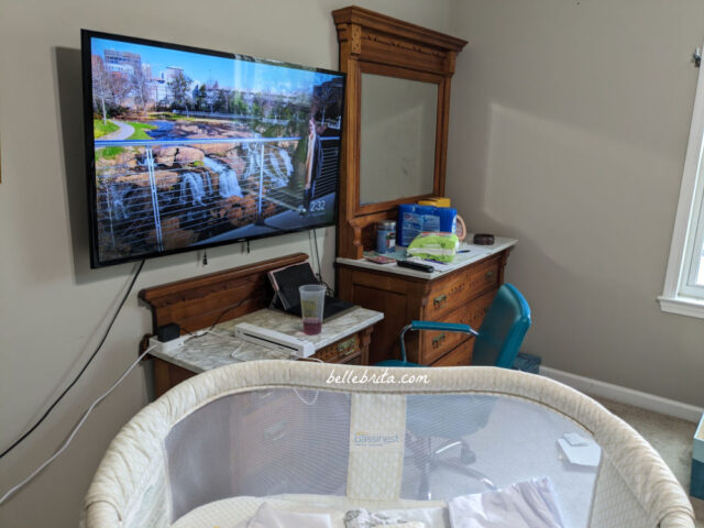 Progress photo of a guest bedroom that will be turned into a nursery. Shows a bassinet, antique wood furniture, and a wall-mounted TV. 
