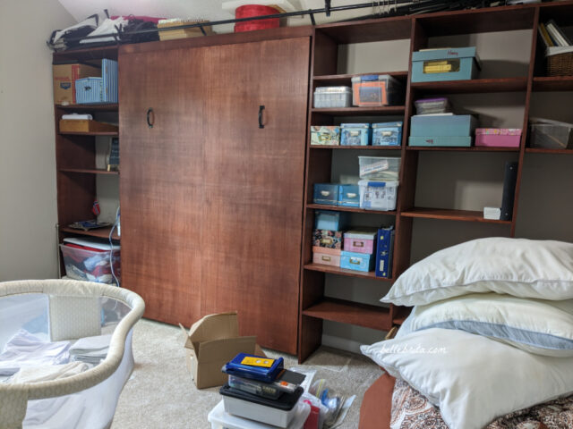 Progress photo of a guest bedroom that will be turned into a nursery. Shows a bassinet, a Murphy bed folded up, shelving with boxes, a stack of bedding and pillows, and other messy things.