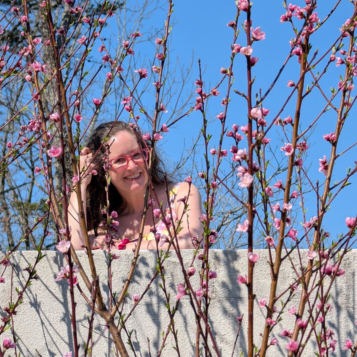 Brita Long, a Millennial white woman with brown hair and hot pink glasses, looks at the camera, smiling. She stands on a castle wall behind peach trees in early bloom with small pink flowers. She is the founder of Belle Brita, a Christian feminist lifestyle blog.