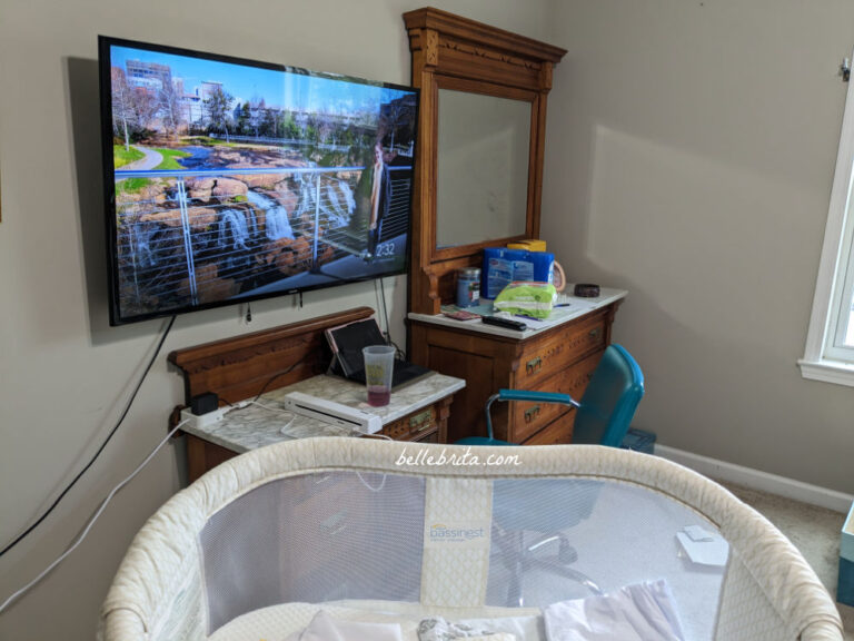Progress photo of a guest bedroom that will be turned into a nursery. Shows a bassinet, antique wood furniture, and a wall-mounted TV.